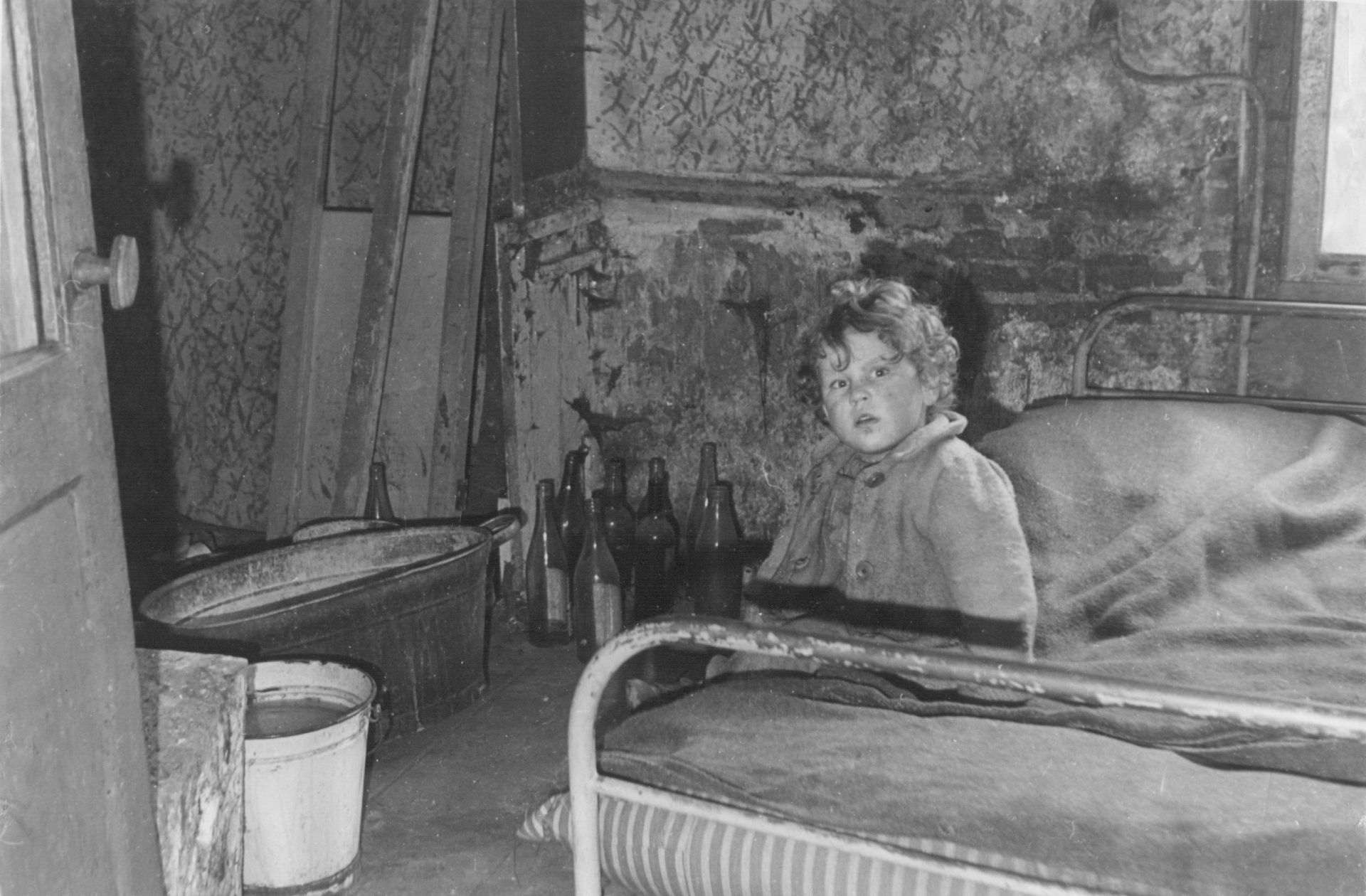 Een kind, kleuter nog zit op het bed in een kamer van een onbewoonbaar verklaarde woning. Er staan drankflessen op de grond.
